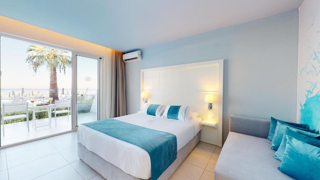 Mūsdienīga viesnīcas numura interjers ar gaišām krāsām un skatu uz jūru. Numurā ir liela gulta, dīvāns un terase ar palmām.