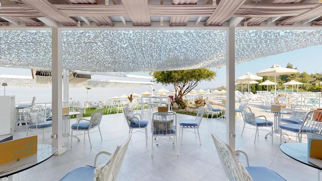 Mūsdienīgs un elegants āra kafejnīcas interjers ar baltām mēbelēm un zilu sēžamvietu, kas piedāvā skatu uz baseinu un pludmali, zem dekoratīva nokrāsojuma.