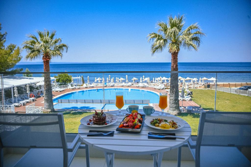 Skats uz baseinu un jūru no terases ar brokastu galdu, kas klāts ar dažādiem ēdieniem un dzērieniem. Palmu koki pie baseina un saulainas pludmales gultas papildina atpūtas atmosfēru.