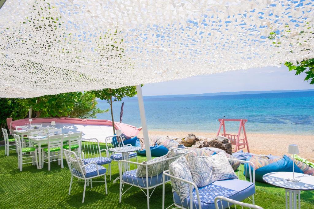 Pludmales restorāna terase ar baltu segumu, zaļiem dārza krēsliem un skatu uz zilo jūru. Ideāla vieta relaksācijai un ēdiena baudīšanai.