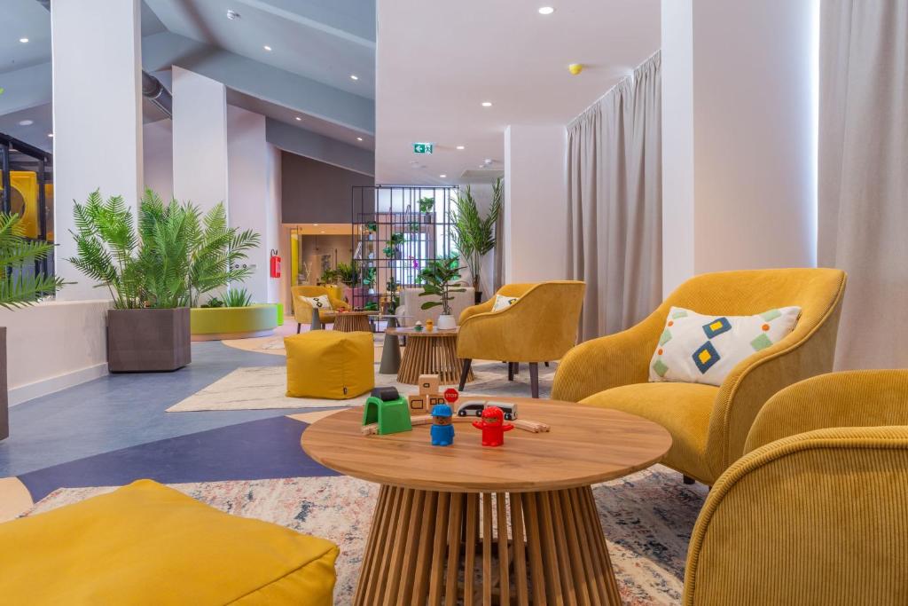 Mūsdienīgs un elegants atpūtas telpas interjers ar dzelteniem krēsliem, koka galdu un rotaļlietu elementiem, veidojot mājīgu un viesmīlīgu atmosfēru. Telpā redzami arī augi un krāsaini sēdvietas.