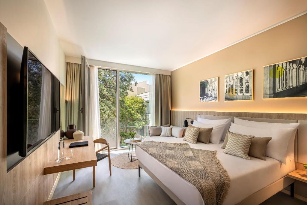 Mūsdienīga viesnīcas numura interjers ar plašu logu, kas sniedz skatu uz dārzu, un modernu televizoru. Gulta ir ērta ar dekoratīvajām spilveniem un tekstilu, radot mājīgu atmosfēru.