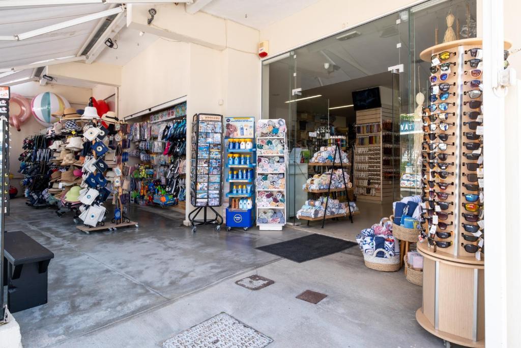 Veikala iekšpuse ar dažādiem piederumiem, tostarp cepurēm, saulesbrillēm un pludmales rotaļlietām. Redzamas krāsainas preces un pārdevēja stends, kas piedāvā tūristu un pludmales aksesuārus.