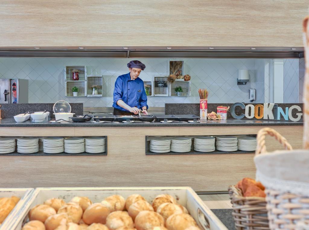 Kulinārijas darbnīca, kur šefpavārs strādā pie plīts, sagatavojot ēdienus. Priekšplānā redzami maizes izstrādājumi, kas papildina virtuves atmosfēru.