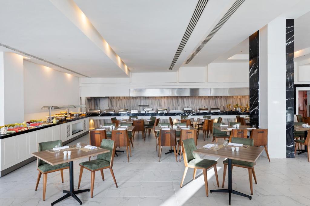 Moderns restorāns ar elegantiem galdiem un krēsliem, kas izvietoti plašā telpā, kurā redzama bufetes zona un gatavošanas zona.