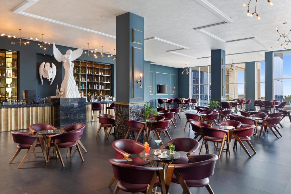 Elegants restorāns ar modernu interjeru, kas ietver stilīgas sarkanās krēslus un baltu skulptūru. Bāra zona ir izsmalcināta ar plašu dzērienu izvēli un gaišām logiem, kas piedāvā skatu uz ārpusi.