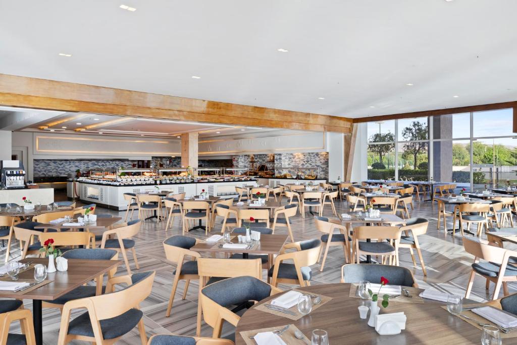 Mūsdienīgs restorāns ar plašu telpu, elegantiem galdiem un krēsliem, kas rotāti ar ziediem. Redzama bāra zona ar ēdienu un dzērienu piedāvājumu, kā arī lieliem logiem, caur kuriem ieplūst dabiska gaisma.