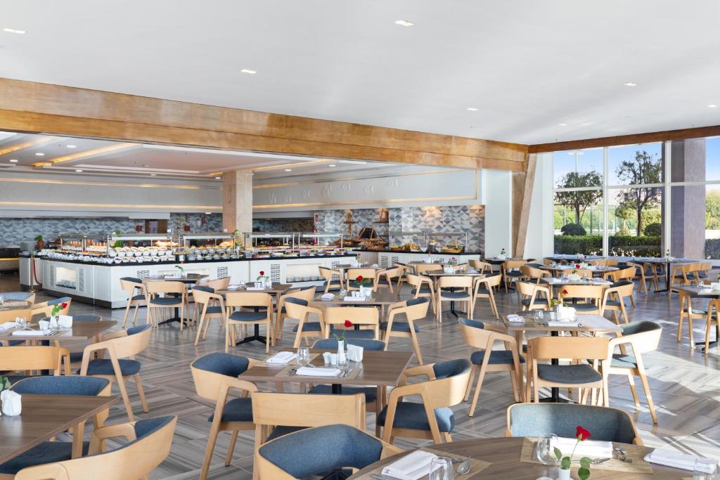 Mūsdienīgs restorāns ar plašu ēdamzāli, kas iekārtots ar koka un zilu krēslu kombināciju. Redzams bufetes galds, kas pilns ar dažādiem ēdieniem, un lieli logi, caur kuriem ieplūst dabiska gaisma.