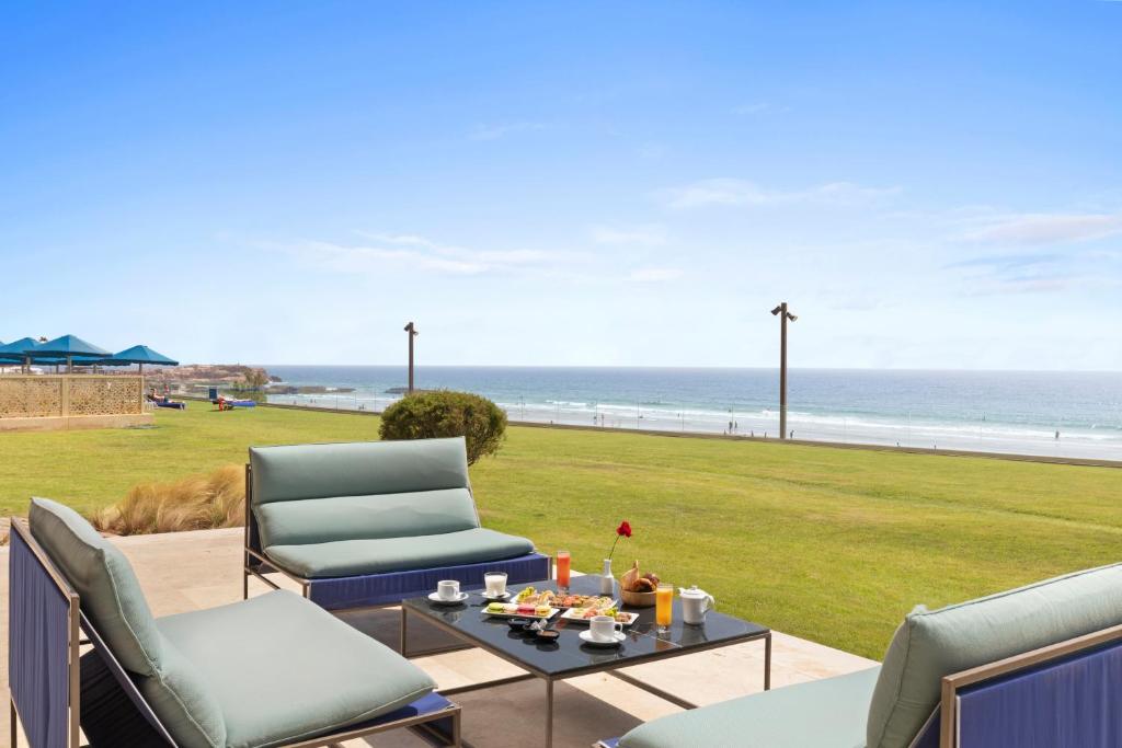 Atpūtas zona ar skatu uz jūru, kurā redzamas ērtas mēbeles un brokastu galds ar dzērieniem un pārtiku. Fons ietver zāles platību un pludmales ainavu.