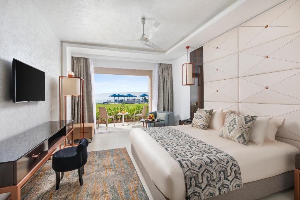 Mūsdienīgs viesnīcas numurs ar plašu logu, kas atveras uz terasi un skatu uz jūru. Numurā ir liela gulta, televizors, eleganta mēbeles un dekoratīvi spilveni.