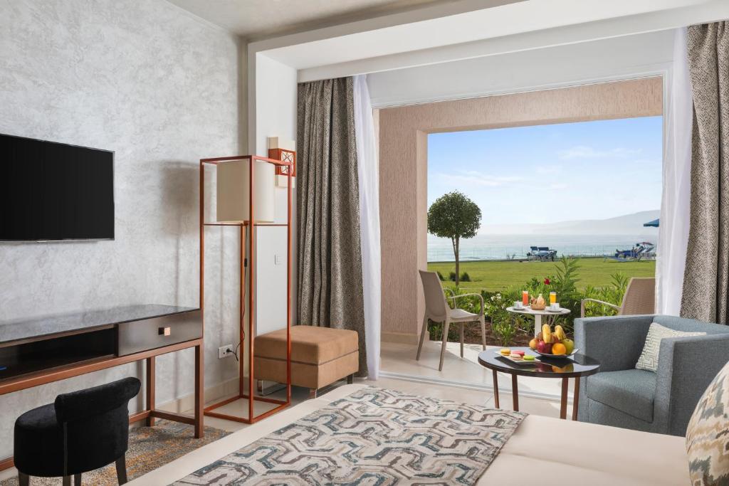 Mūsdienīga viesnīcas istaba ar plašu skatu uz jūru, kurā redzami komfortabli mēbeļu elementi un dzīvžogs. Loga tuvumā ir galdiņš ar augļiem un dzērieniem.