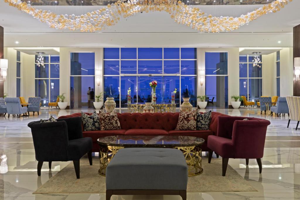 Mūsdienīgs viesnīcas vestibilis ar elegantu sarkanu dīvānu, stikla galdu un dažādu krāsu krēsliem. Gaismas elementi un plašas logu ailes rada patīkamu atmosfēru.