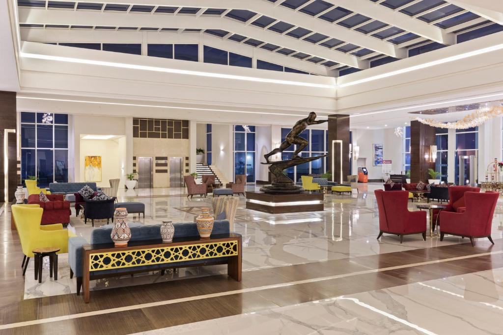 Mūsdienīgs viesnīcas vestibils ar elegantu dizainu, kurā ir dažādas krāsainas mēbeles un skulptūra centrā. Telpā ir lieli logi, kas nodrošina dabisko apgaismojumu un plašuma sajūtu.