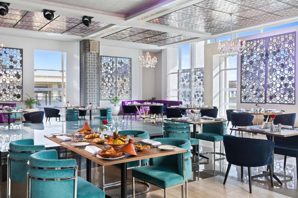 Elegants restorāna interjers ar modernām mēbelēm, plašām logu atverēm un dekoratīviem elementiem. Uz galda izvietoti ēdieni, radot viesmīlīgu atmosfēru.