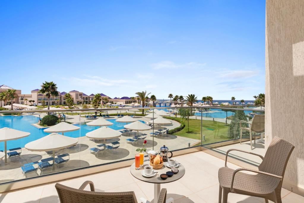 Balkona skats uz luksusa viesnīcas baseinu un atpūtas zonu, kurā redzami sauļošanās krēsli un palmas. Uz galda izvietota brokastu komplekts ar dzērieniem un uzkodām.