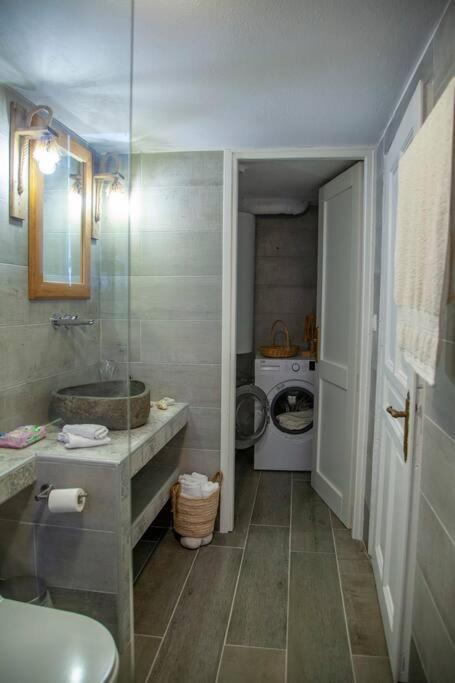 Mūsdienīgs vannas istabas interjers ar akmens izlietni, stikla dušu un veļas mašīnu. Telpa ir gaiša un eleganta, ar pelēkām flīzēm un koka akcentiem.