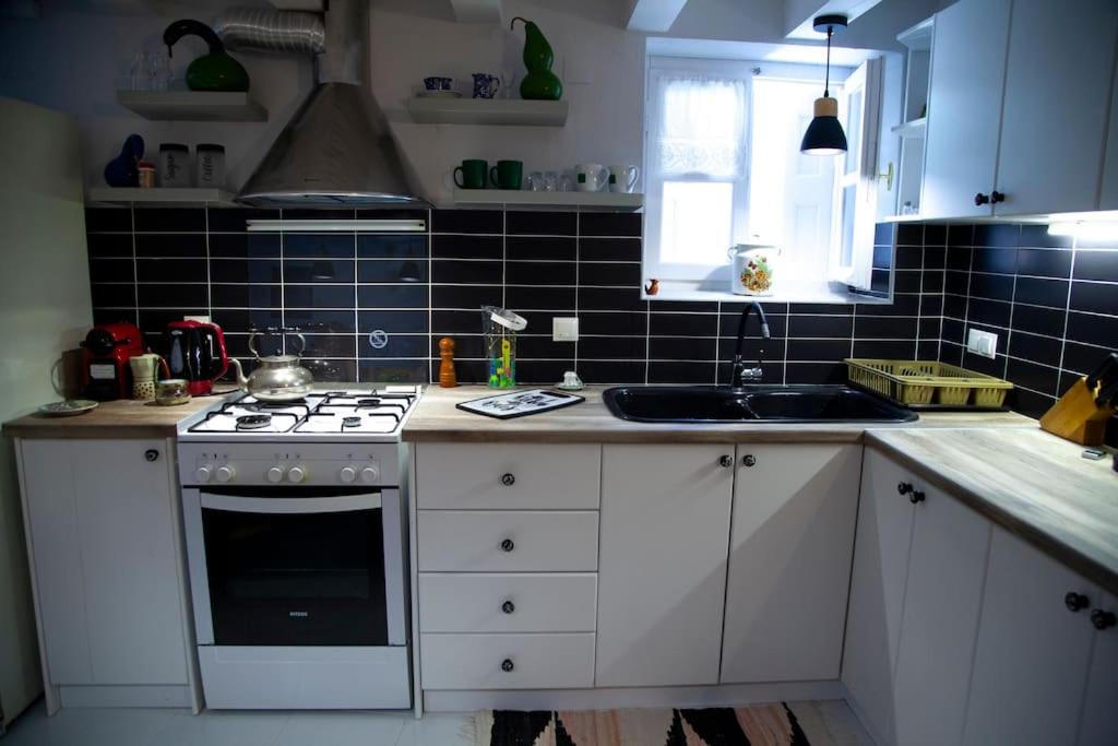 Mūsdienīga virtuve ar melnu flīžu sienu un baltām skapīša durvīm, kurā atrodas gāzes plīts, izlietne un dažādi virtuves piederumi. Interjers ir ērts un funkcionāls, radot patīkamu atmosfēru.