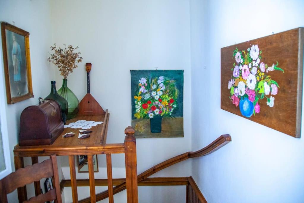 Mājīgā kāpņu telpa ar koka margām, krāsainām ziedu gleznām uz sienām un vintage priekšmetiem uz galda, radot siltu un mājīgu atmosfēru.