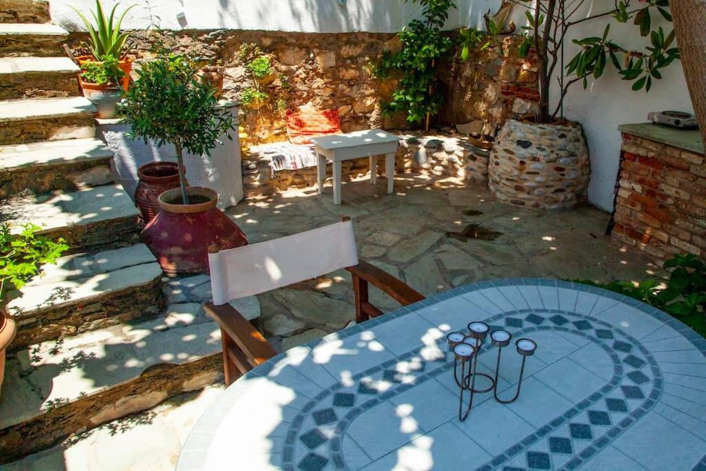 Skaista dārza telpa ar akmens celiņu, ziediem un mēbelēm, kas nodrošina mierīgu un relaksējošu atmosfēru. Uz galda atrodas sveču turētājs, kas piešķir šarmu šai vietai.