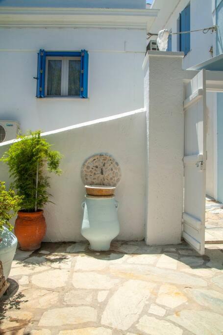 Skaista, saulaina iekštelpa ar akmens grīdu, kurā redzams balts pods ar koka vāku un akmens detaļu, blakus zaļai auga podiņam un sarkaniem podiem.