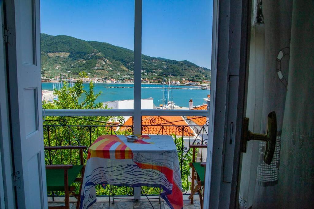 Skats no balkona uz jūru ar laivām un kalniem fona. Uz galda ir krāsains galdauts un trauks, radot mājīgu atmosfēru.