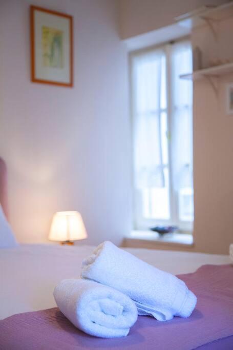 Mīksti, balti dvieļi novietoti uz gultas, blakus nelielai lampiņai un logam, kas piepilda telpu ar dabiskiem gaismas stariem.