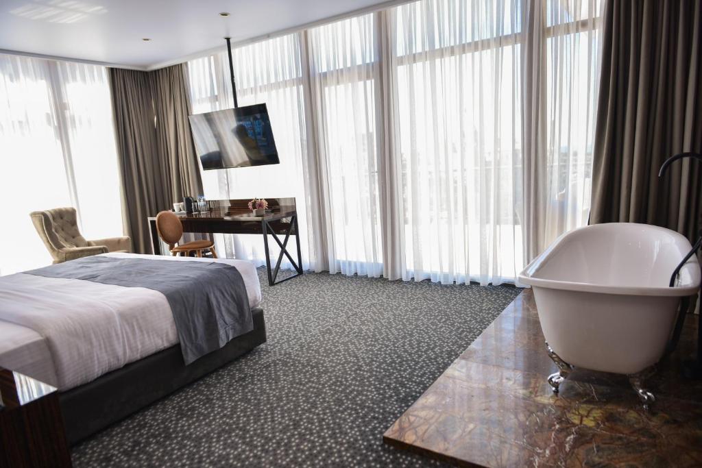 Mūsdienīga viesnīcas istaba ar plašu logu, elegantu gultu, rakstāmgaldu un brīvstāvošu vannu. Gaisīga un komfortabla atmosfēra.
