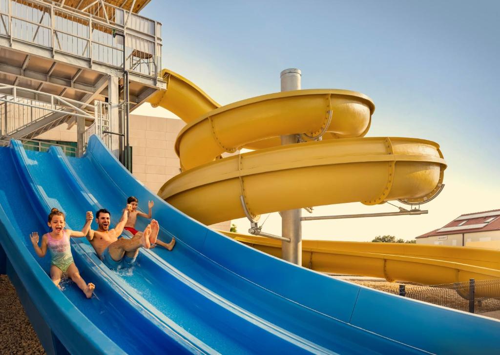 Ūdens slidkalniņš ar trīs personām, kas priecīgi slīd pa zilo virsmu, izbaudot vasaras izklaidi. Fons ar dzeltena krāsas slidkalniņu un skaidru debesu.