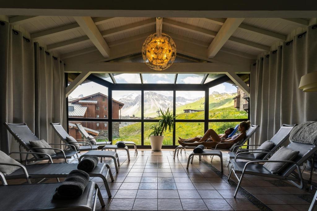 Relaksējoša atpūtas telpa ar skatu uz kalniem, kur divi cilvēki atpūšas krēslos. Telpā ir daudz gaismas un zaļumi, kas radījuši mierīgu atmosfēru.