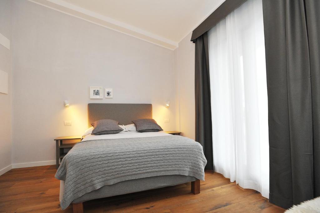 Mūsdienīgs guļamistabas interjers ar pelēku gultu un dekoratīvām spilveniem, apgaismots ar stilīgām sienas lampām. Labi iekārtota telpa ar koka grīdu un elegantām aizkariem.