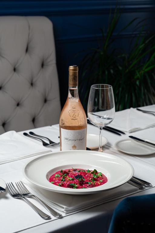 Eleganta maltīte ar koši sarkano ēdienu un pudeli rozā vīna uz galda, ko apņem baltas galdautas un eleganta galda piederumi.