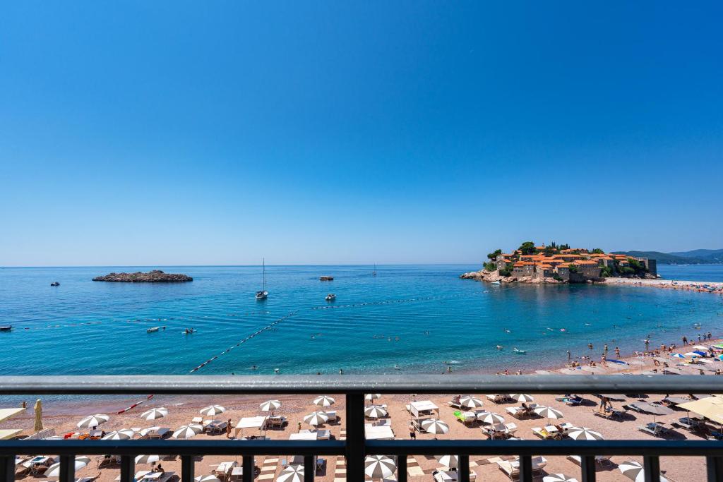 Skaists skats uz jūru ar pludmales zonu, kur redzami sauļošanās krēsli un lietussargi, kā arī neliela sala ar ēkām fona. Jūra ir mierīga un dzidra, ar dažām laivām un peldētājiem.