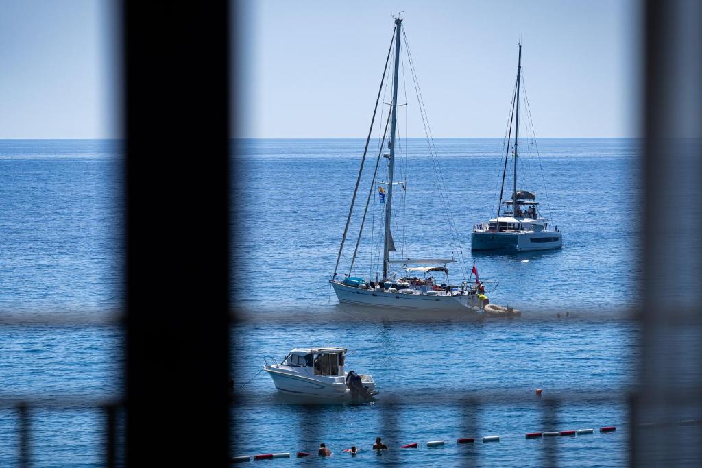 Trīs laivas mierīgā jūrā, redzamas caur žogu. Zilas debesis un saulaina vide rada relaksējošu atmosfēru.
