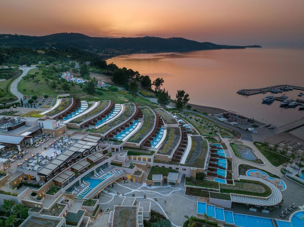 Vistas nozares skats uz modernu kūrortu, kas atrodas pie jūras, ar baseiniem un dārziem, kas saplūst ar ainavu, un saulriets, kas izgaismo debesu krāsas.