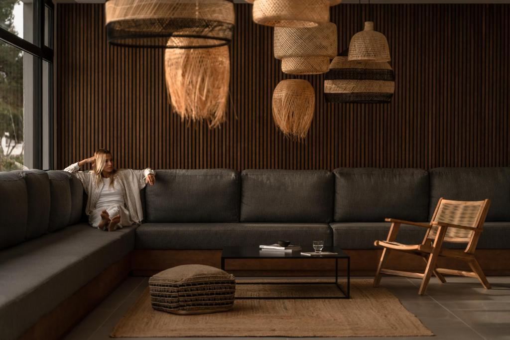 Mūsdienīga viesistaba ar pelēku dīvānu un stilīgiem, rotājošiem lampām, kurā atpūšas sieviete. Telpa ir iekārtota ar dabīgiem materiāliem un siltām krāsām, radot mierīgu atmosfēru.