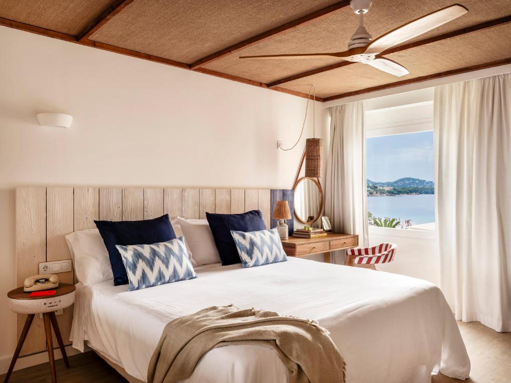 Mūsdienīgs guļamistabas interjers ar baltu gultu un dekoratīvām spilveniem, koka galdu un skatu uz jūru caur logu. Eleganta un relaksējoša atmosfēra.