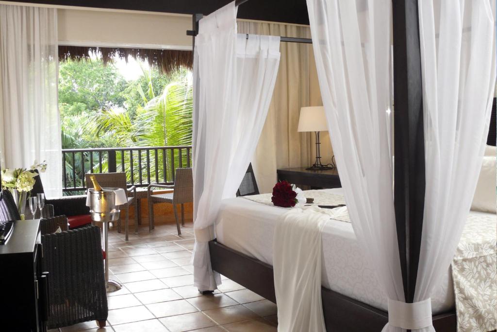 Elegants guļamistabas interjers ar četrstūra gultu, apņemtu ar vieglām, baltām aizkariem. Logi veras uz zaļu dārzu, sniedzot mierīgu atmosfēru. Galds ar lampu un sēdvietas pie loga papildina telpas komfortu.