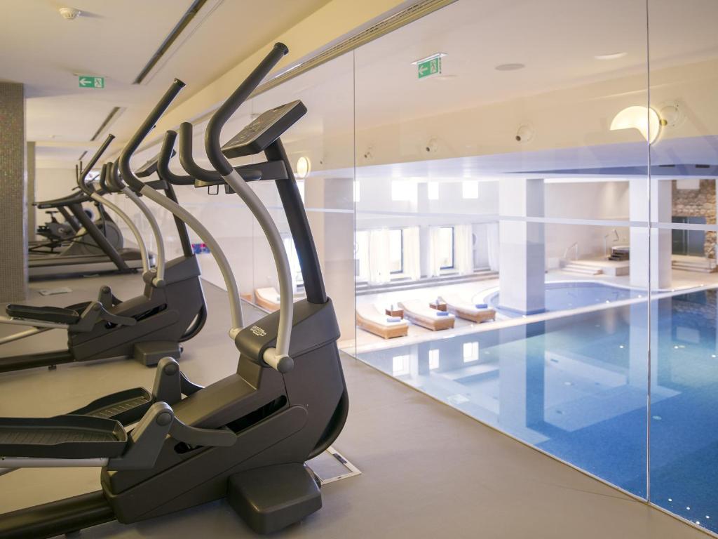 Mūsdienīgs sporta zāles iekārtojums ar eliptiskajiem trenažieriem un skatu uz iekštelpu baseinu. Gaismas un minimālisma dizains veido relaksējošu atmosfēru treniņiem.