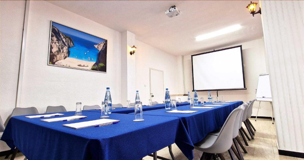 Konferenču telpa ar zilu galdu, ūdens pudelēm un piezīmju lapām. Sienā ir gleznains pludmales attēls, un telpā ir projektors un ekrāns.