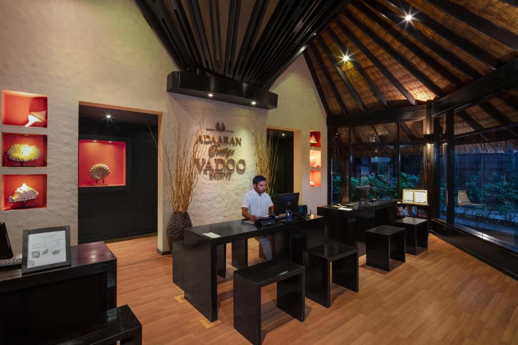 Viesnīcas reģistratūra ar modernu dizainu un bambusa jumtu, kur darbinieks apkalpo viesus. Interjerā ir siltas krāsas un dekoratīvi elementi, kas rada viesmīlīgu atmosfēru.