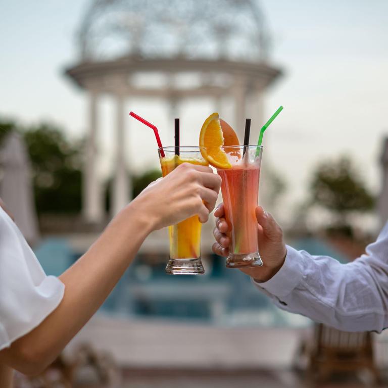 Divi cilvēki, satiekoties ar kokteiļiem, svin brīvdienas, baudot dzērienus ar apelsīnu sagriezumiem, fonā redzama eleganta terasa un baseins.