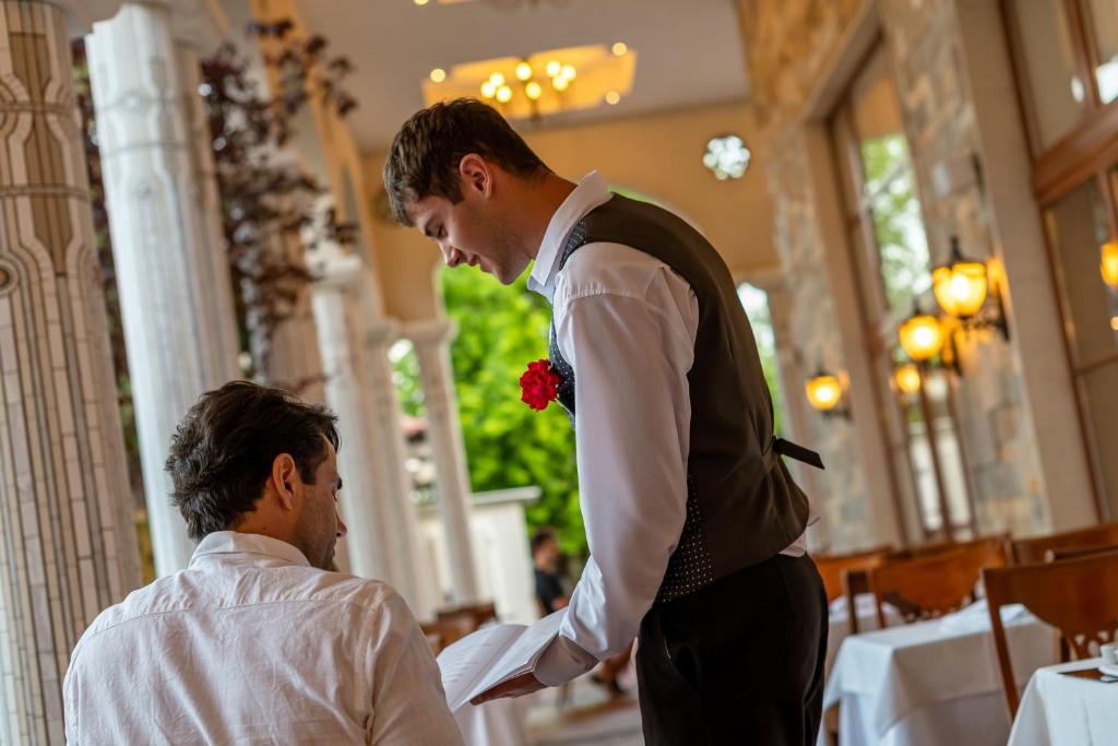 Viesmīlis pasniedz ēdienkarti klientam eleganta restorāna vidē. Gaisotne ir mājīga un relaksējoša, ar dekoratīvām gaismām un stilīgiem interjera elementiem.