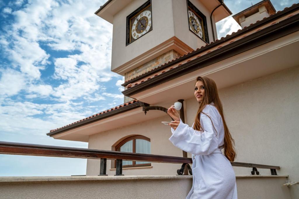 Sieviete baltā halātā tur kafijas krūzi uz balkona, skatoties uz debesīm ar mākoņiem un pulksteņa torni fonā.