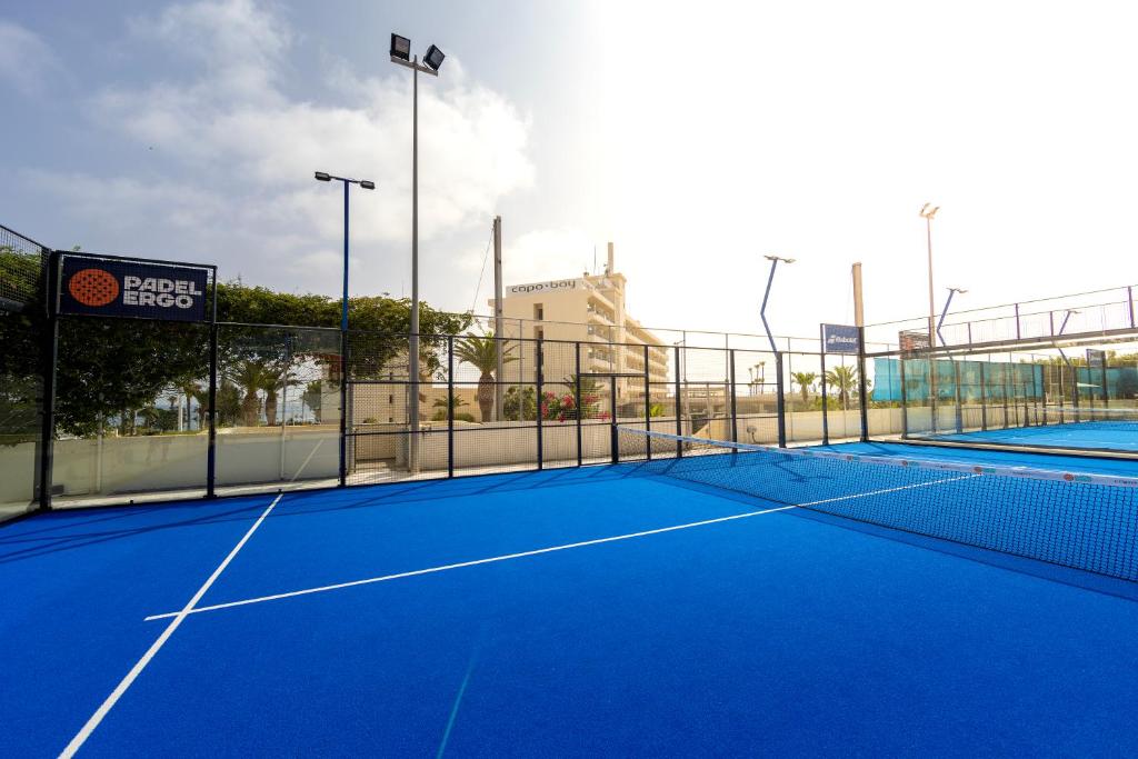 Zilā padel tenisa laukuma skats ar zīmolu "Padel Ergo" un apgaismojuma stabiem. Fons attēlo ēkas un zaļus augus, radot patīkamu atmosfēru sportošanai.