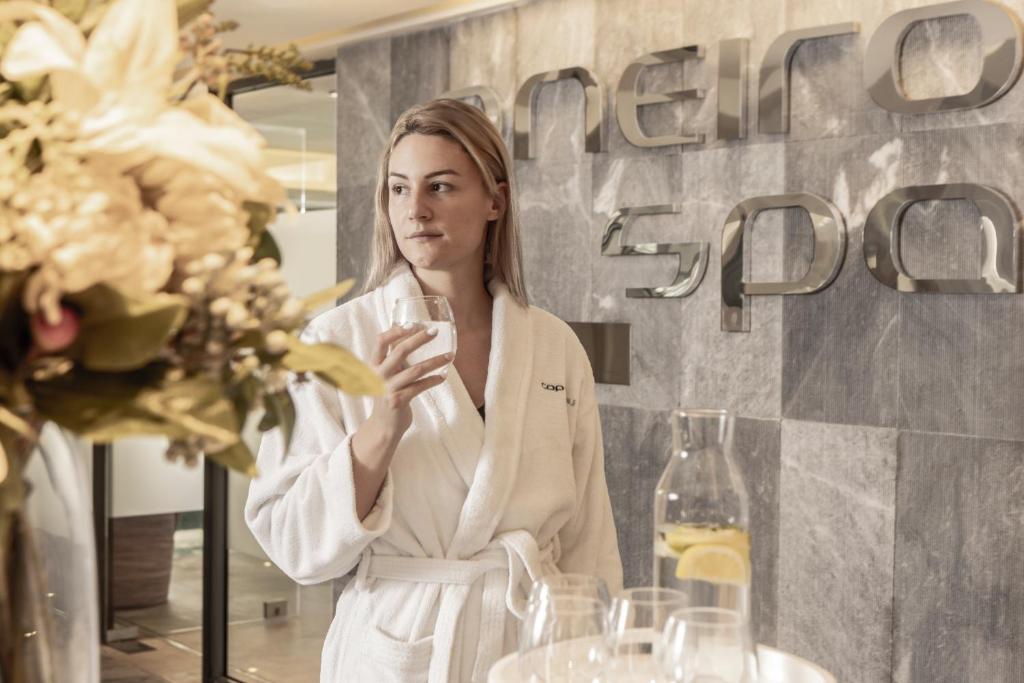 Sieviete baltā halātā, turot glāzi ūdens, stāv pie modernā spa logo fona, kas izveidots no spīdīgas metāla. Blakus ir ziedu kompozīcija un ūdens karafes ar citrona šķēlēm.
