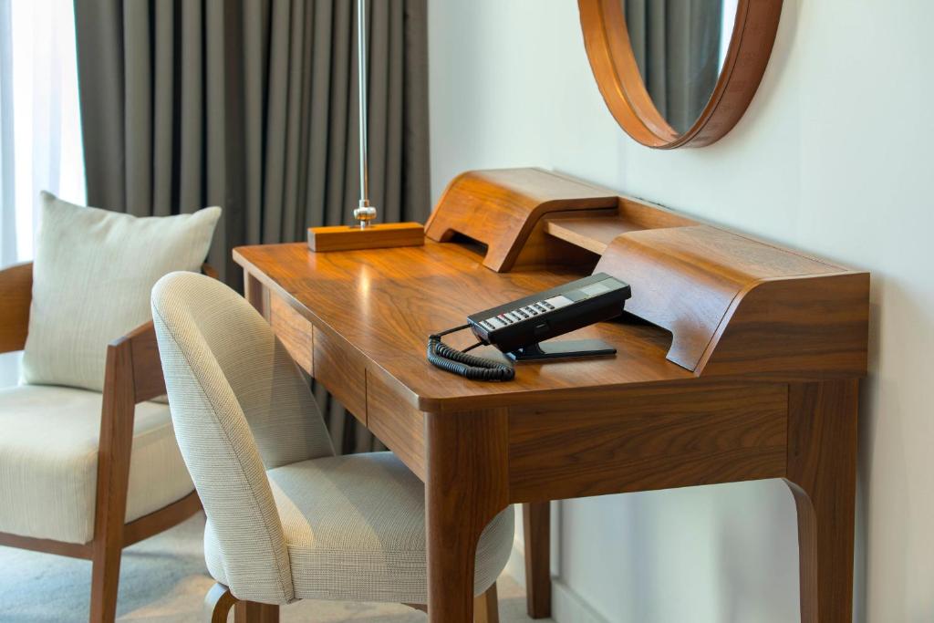 Mūsdienīgs darba galds ar koka apdari, telefona aparātu un ērtu krēslu, blakus atpūtas krēslam ar spilvenu. Interjers ir gaišs ar elegantām aizkariem un apaļu spoguli uz sienas.
