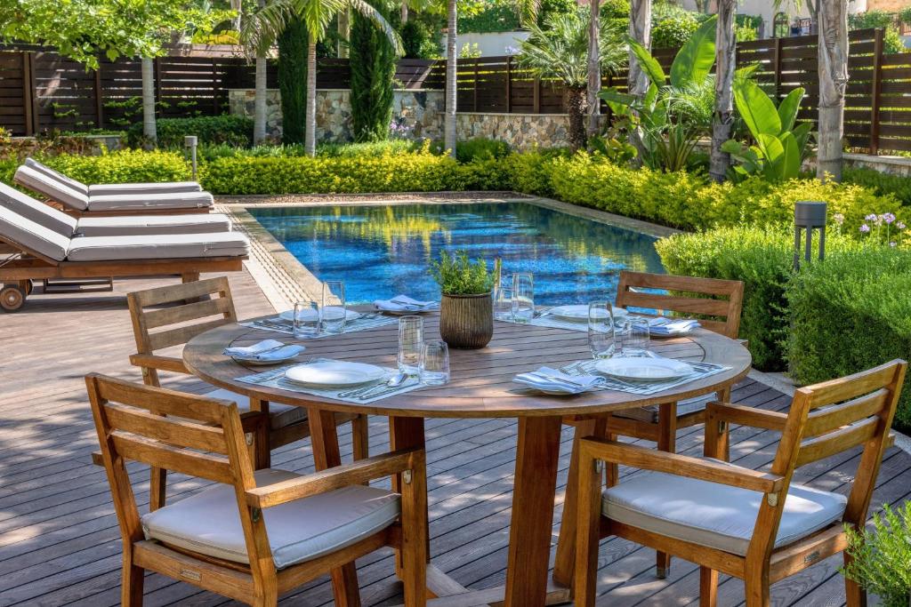 Āra ēdamgalds ar koka krēsliem un galdu, ap kuru saklāti galda piederumi, blakus baseinam un sauļošanās krēsliem, ieskauts zaļo augu ainavā.