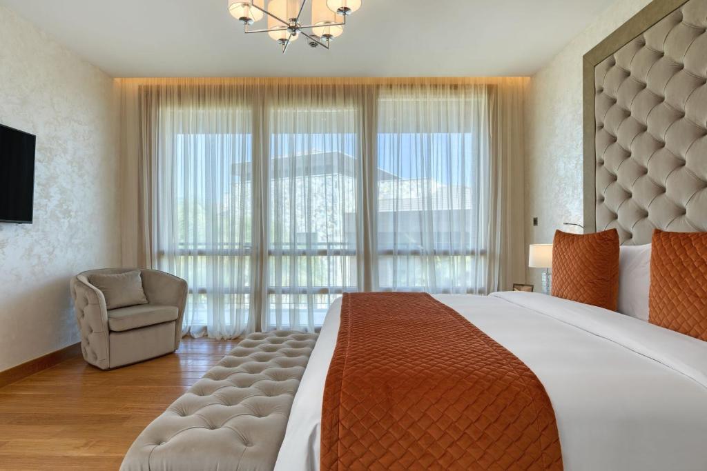 Mūsdienīgs guļamistabas interjers ar elegantu gultu un oranžiem spilveniem, liela loga ar caurspīdīgām aizkariem un komfortablu atpūtas krēslu.