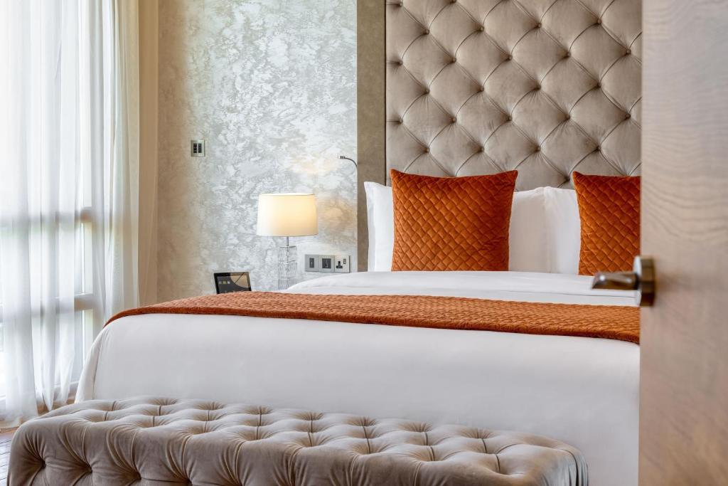 Elegants guļamistabas interjers ar krēmkrāsas sienām un greznu gultu, kas rotāta ar spilveniem ogļu krāsā. Blakus gultai atrodas moderna lampa un ērta mīksta krēsla.