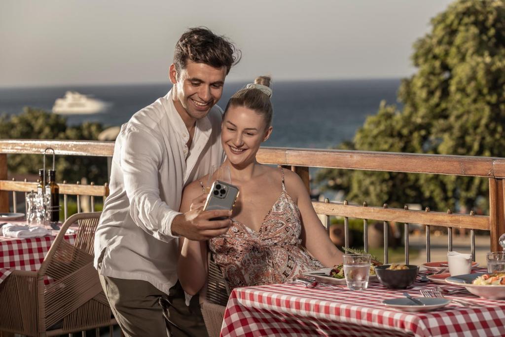 Pāris sēž restorānā ar skatu uz jūru, priecīgi skatoties telefonu. Uz galda ir ēdiens un dzērieni, radot patīkamu atmosfēru.